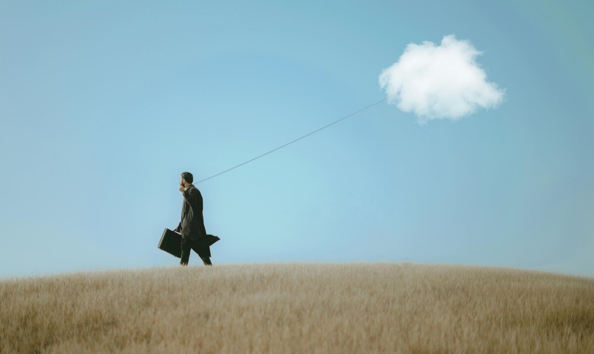 Man walking through golden wheat field tethered to floating white cloud by invisible string