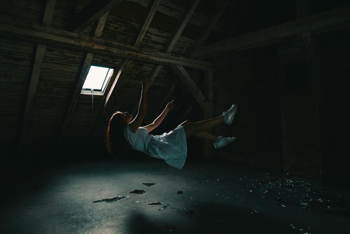 Woman levitating horizontally in dark abandoned attic illuminated by single skylight beam