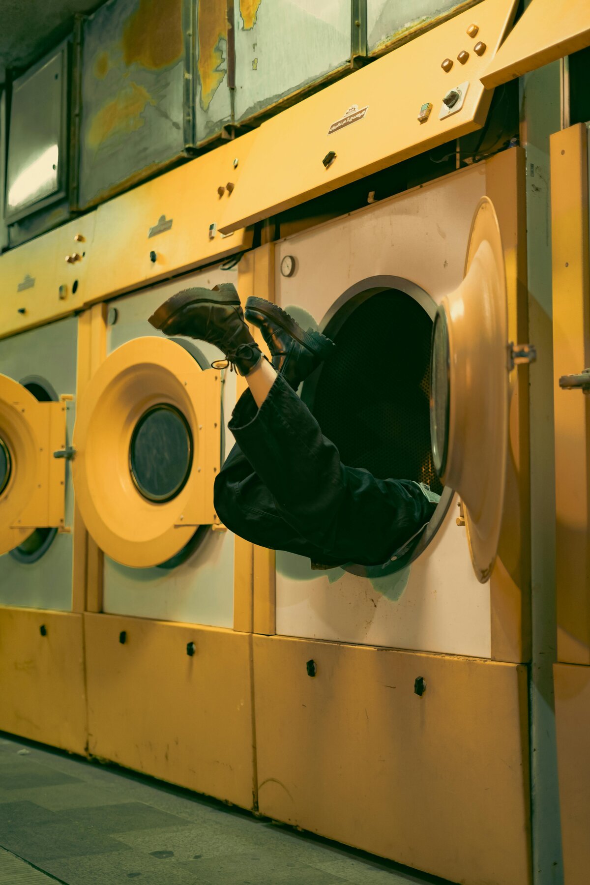 Person climbing into vintage yellow washing machine drum in retro laundromat creating impossible perspective